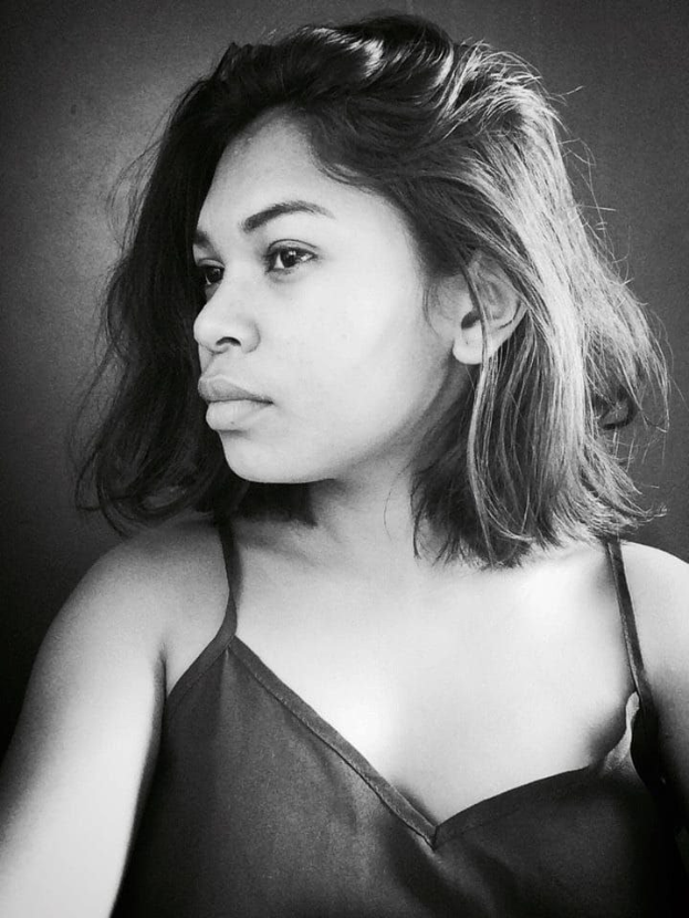 Eine selbstbewusste Frau in einem schwarzen Kleid posiert in einem Schwarz-Weiß-Foto, ihr glatter Bob und ihr dezentes Make-up unterstreichen ihre selbstbewusste Haltung vor einer neutralen Wand.