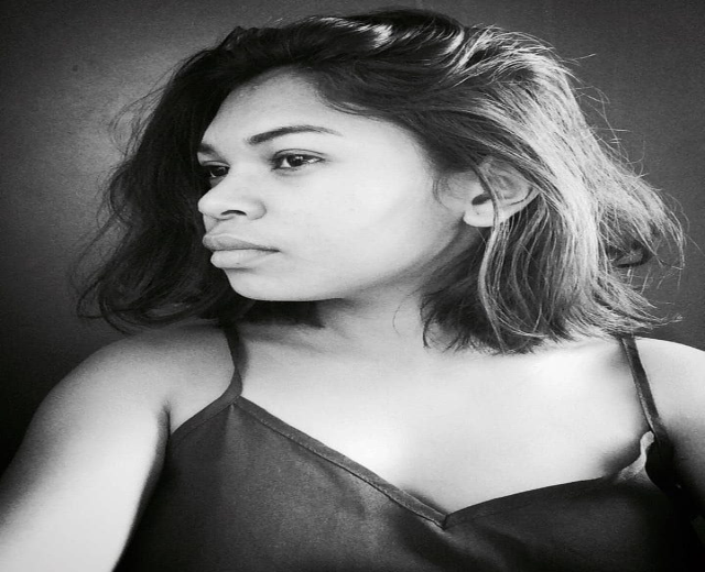 Eine selbstbewusste Frau in einem schwarzen Kleid posiert in einem Schwarz-Weiß-Foto, ihr glatter Bob und ihr dezentes Make-up unterstreichen ihre selbstbewusste Haltung vor einer neutralen Wand.