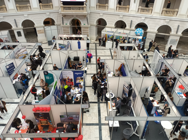 Eine große Gruppe von Menschen auf einer internationalen Messe mit Ständen, Bannern, Tischen, Stühlen, Luftballons und einem Gebäude mit Bögen, Säulen, Geländern und einem Hintergrundbild.