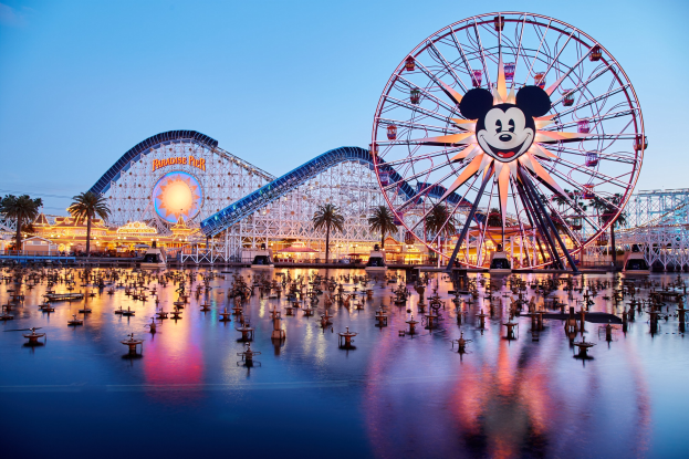 Eine lebendige Disneyland California Adventure-Szene bei Sonnenuntergang mit einem riesigen Mickey-Maus-Rad im Vordergrund, einer Achterbahn im Hintergrund und Booten auf dem Wasser, mit einem Himmel aus Orangen, Pinks und Purpurs und silhouettenartigen Bäumen.