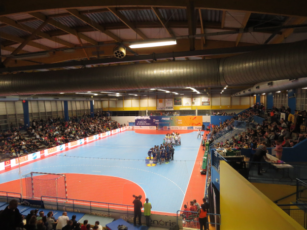 Eine große Halle mit Zuschauern bei einem Handballspiel, mit Sitzplätzen und Stehplätzen, einem Netz auf der linken Seite, Hintergrundtafeln mit Text und Deckenleuchten und Rohren.