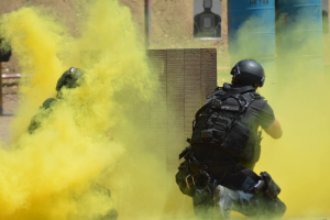 Zwei Polizisten in Einsatzausrüstung mit gelbem Rauch aus dem Mund, vor einer Wand mit einer Tafel und anderen Gegenständen stehend.