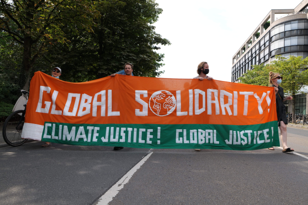 Gruppe von Menschen, die eine Straße entlanggehen und eine "Globale Solidarität: Klimagerechtigkeit"-Plakette halten, mit Fahrrädern, die an der Seite geparkt sind und Gebäuden im Hintergrund.